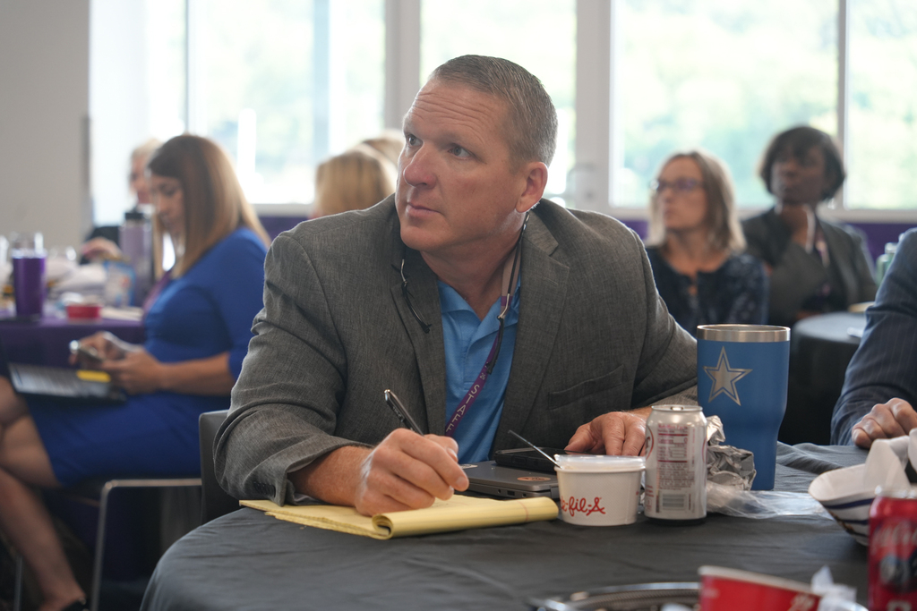 A man in a blue shirt and gray blazer sits at a table with a cup, can, and pen.
