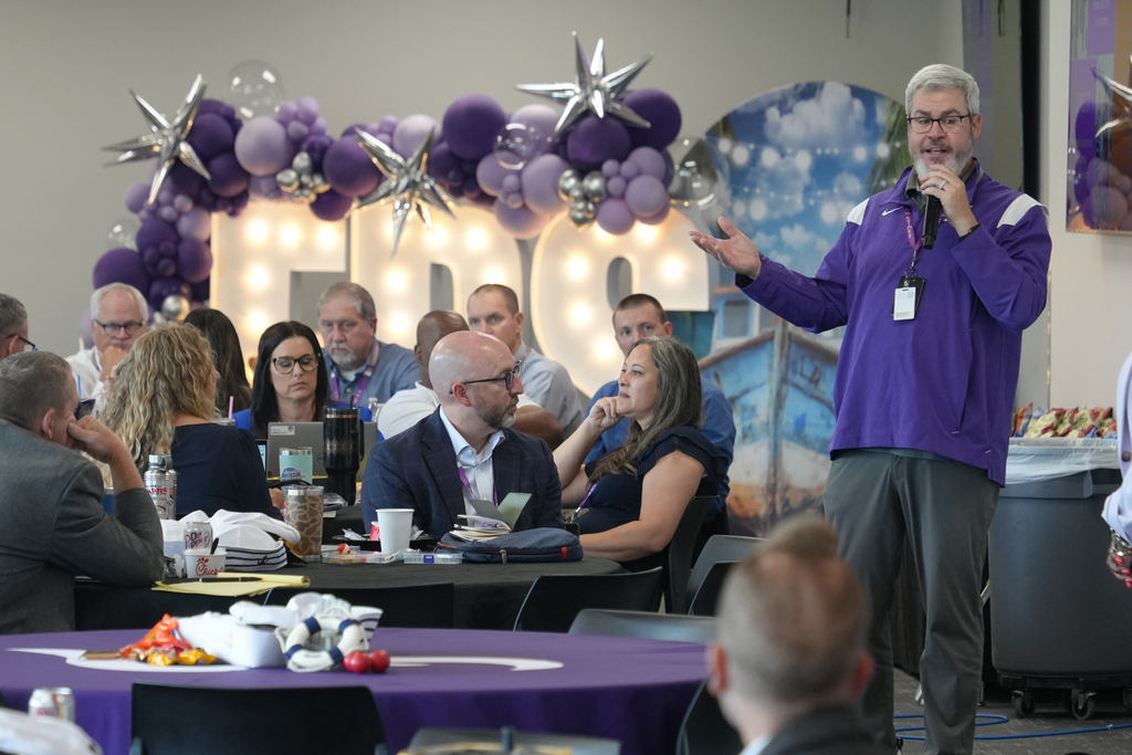 A man speaks into a microphone at a seated event with purple tablecloths and balloons.