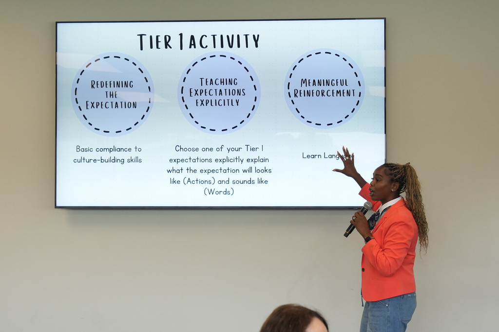 Woman speaking with a microphone in front of a large screen with a presentation on "Tier 1 Activity."