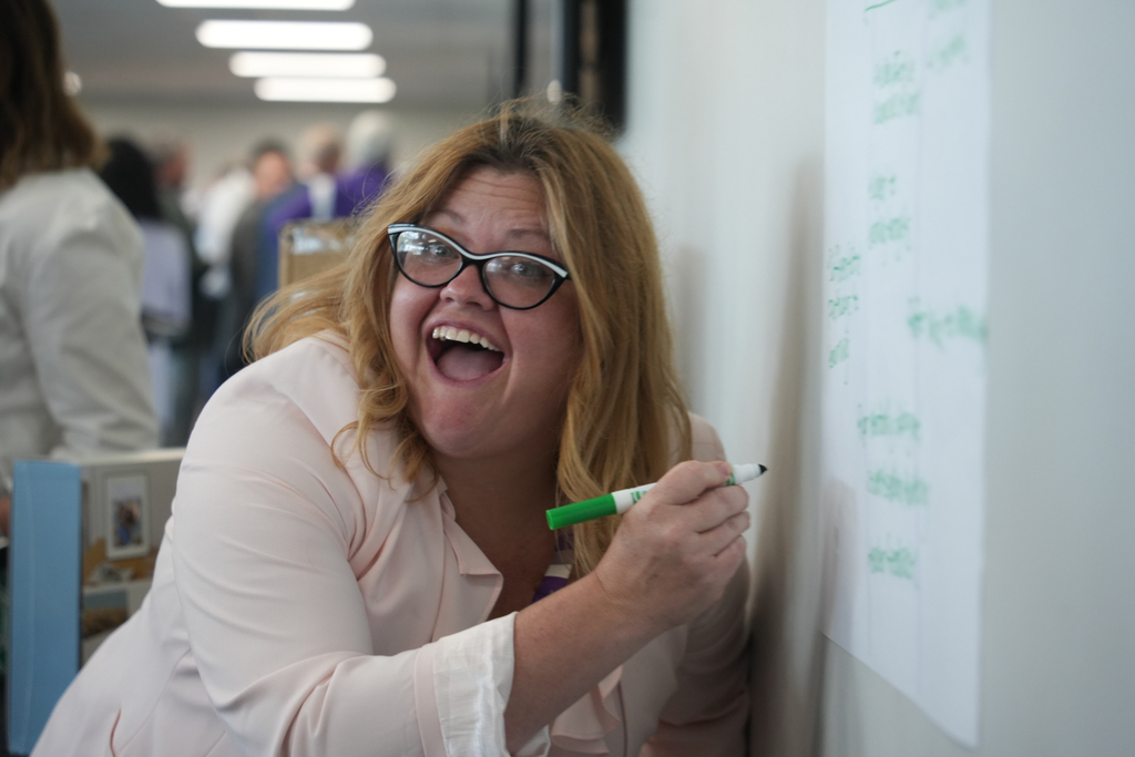 A woman with glasses and blonde hair leans over a white wall, writing on a whiteboard with a green marker.