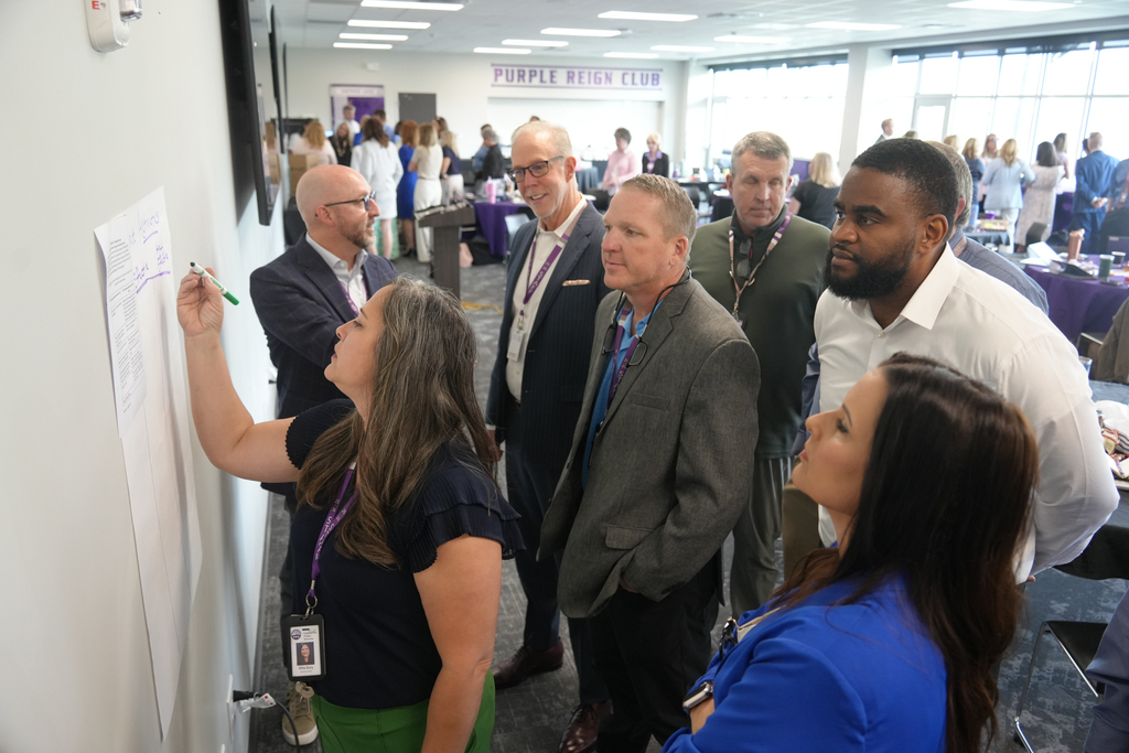 A group of professionals gathered, some writing on a whiteboard, others observing. A woman with green lanyard stands writing.