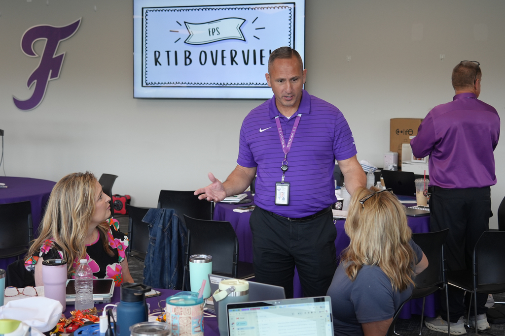 A man in a purple shirt giving a presentation stands before three women seated at a table.