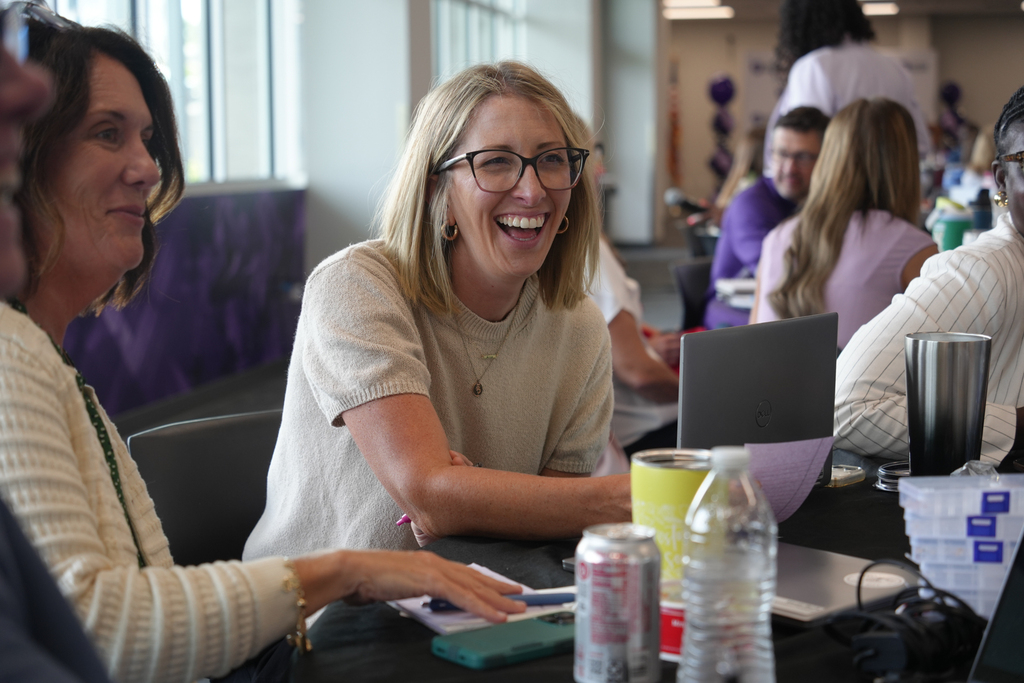 People sit at a table with laptops, cups, and bottles, with one woman smiling and gesturing.