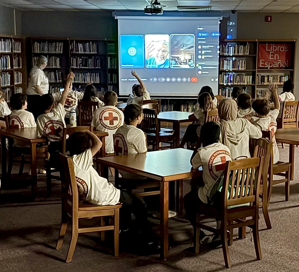 A classroom with students seated around tables; a teacher stands near a projector screen displaying a presentation.