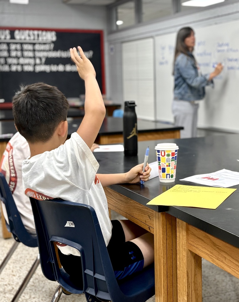 A classroom setting with a boy raising his hand and another person writing on a whiteboard.