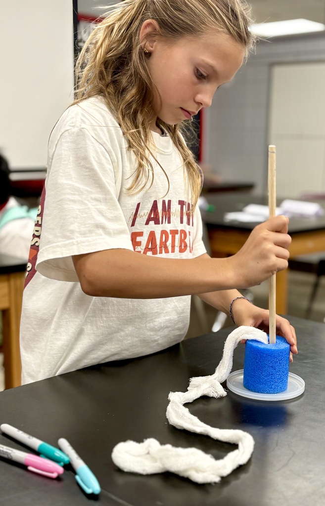 A girl wearing a white T-shirt uses a stick to create yarn around a blue cylinder.