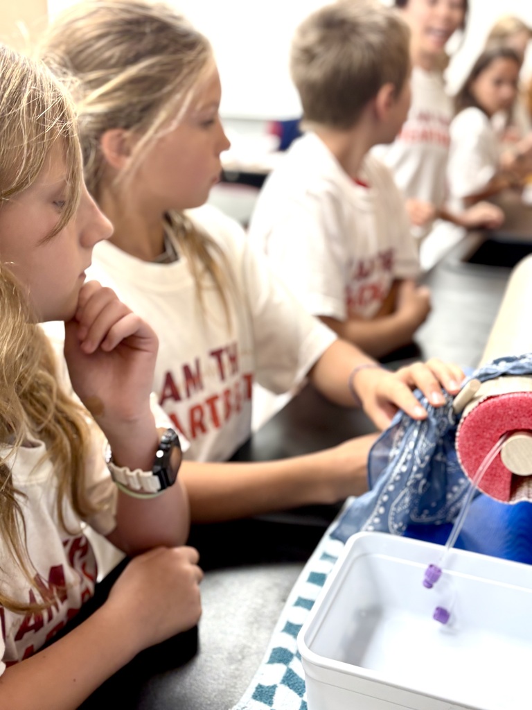 Children in white shirts seated in a classroom, one child holds an art project.