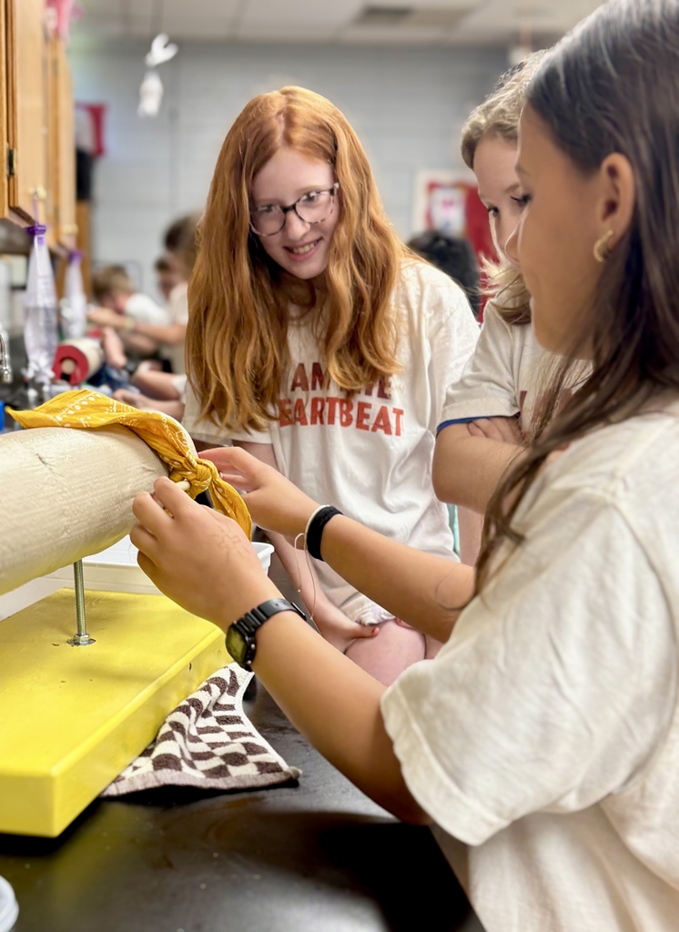 Three students in a classroom setting. One is holding a cloth while another adjusts a yellow device.