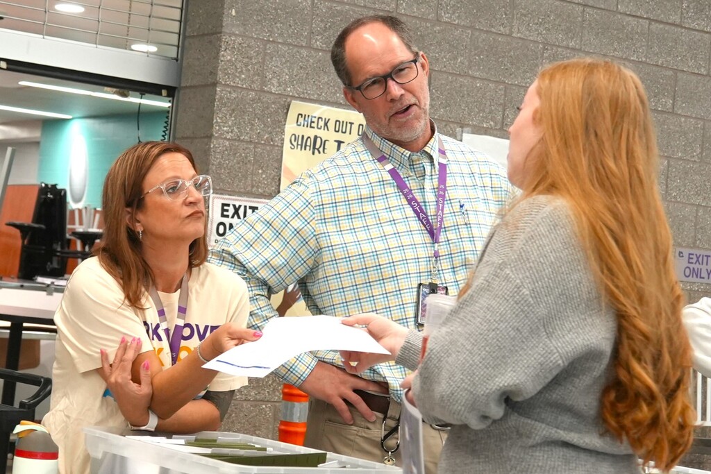 Three adults stand around a table. A man hands a paper to a woman while another woman watches.