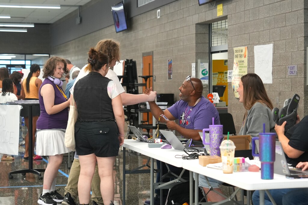 People standing around tables at a gathering, with a person in a purple shirt shaking hands with another.