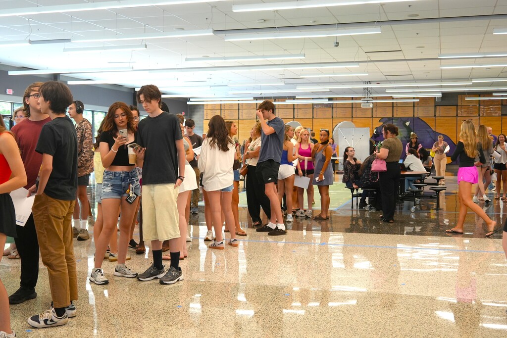People in a hallway, some standing and some sitting. They wear casual attire; some hold papers. Bright lighting.