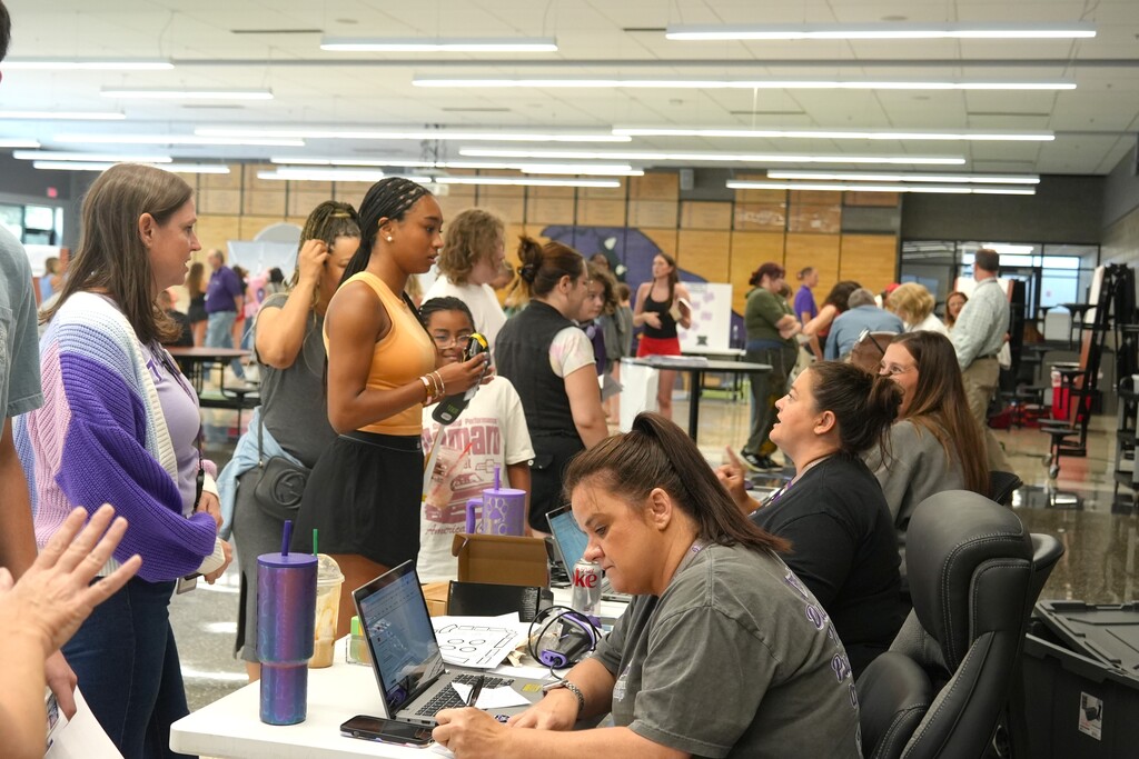 A group of people stand around tables with laptops and materials in an indoor space.