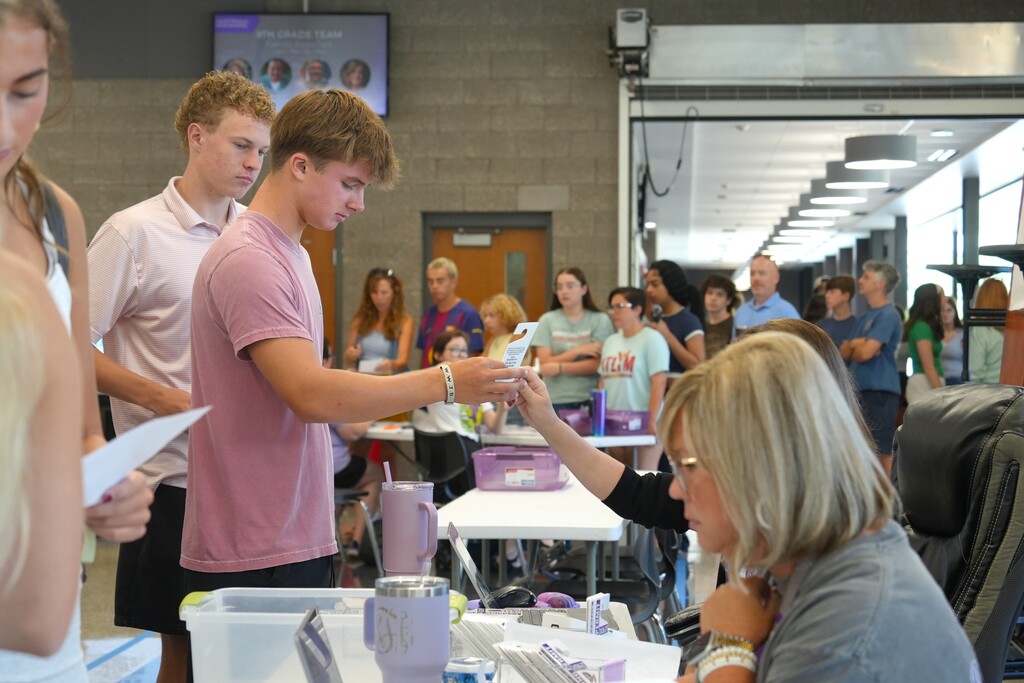 People gather in a room with tables and chairs. A man in a pink shirt and a woman in a gray shirt interact.