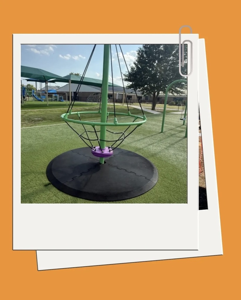 Circular playground equipment with green frame and purple center on a green field with a tree and buildings in the background.