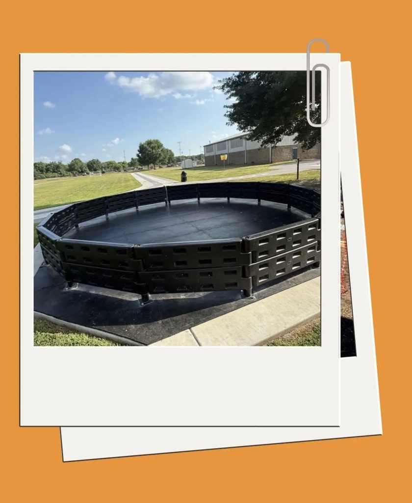 Black circular structure on grass. A building and trees are in the background. Orange background.