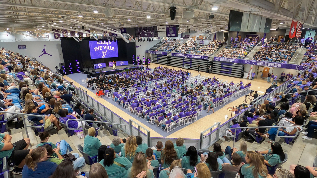 A large crowd fills the stands of an indoor arena. Many attendees wear purple shirts.