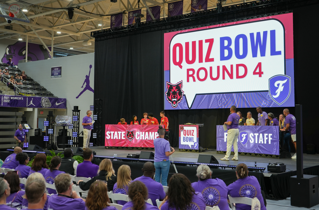 An indoor stadium hosts a quiz bowl round four. Participants stand at a podium. The audience, all in purple, listens.