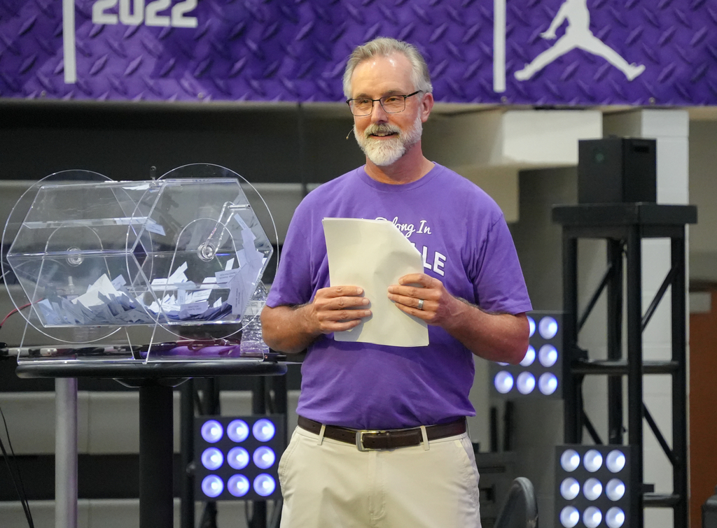A man stands holding papers, wearing a purple shirt, next to a clear box with crumpled paper inside.