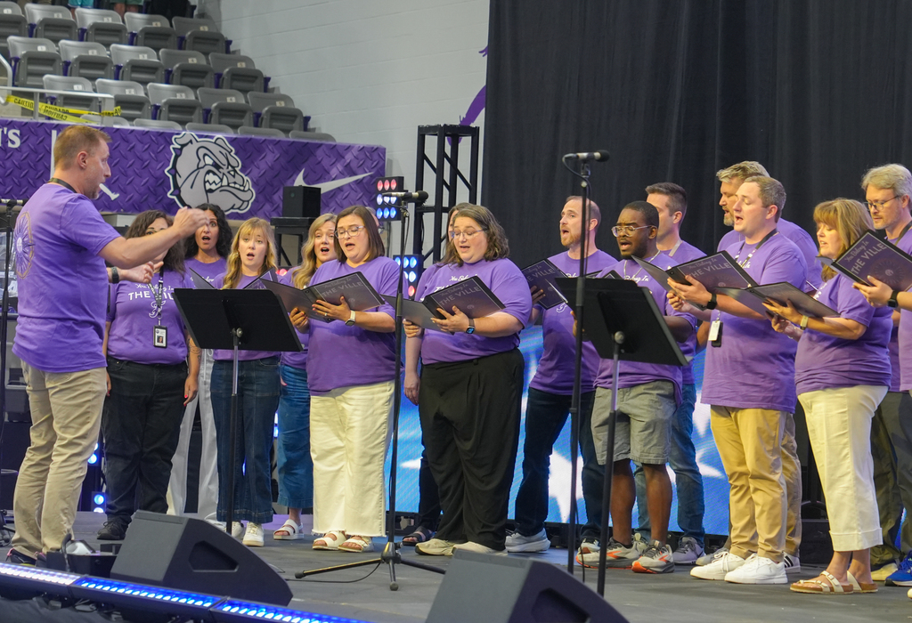 A choir performs on stage with purple shirts. A man stands in front. Microphones, speakers, and music sheets.