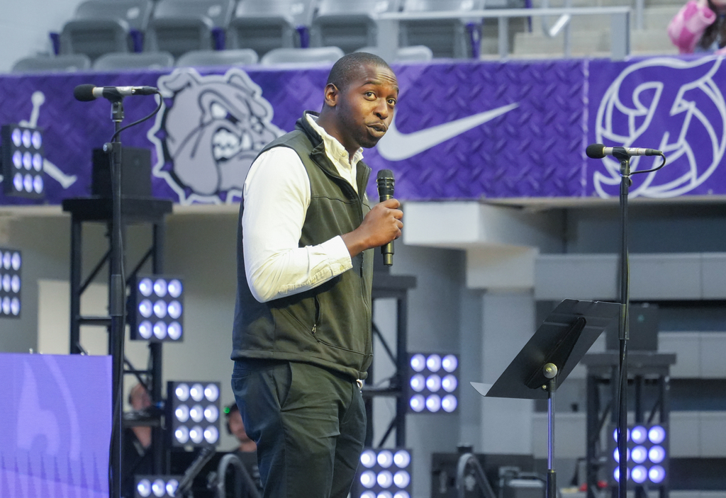 A man stands on a stage, addressing an audience, with microphones and a purple backdrop behind him.