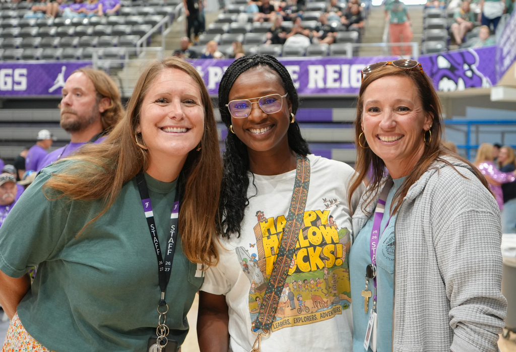 Three women smile for the camera at an event; one wears glasses and a white shirt with a colorful design.