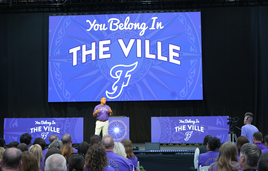A man speaks on stage with a large screen behind him. A large audience sits in front of the stage.