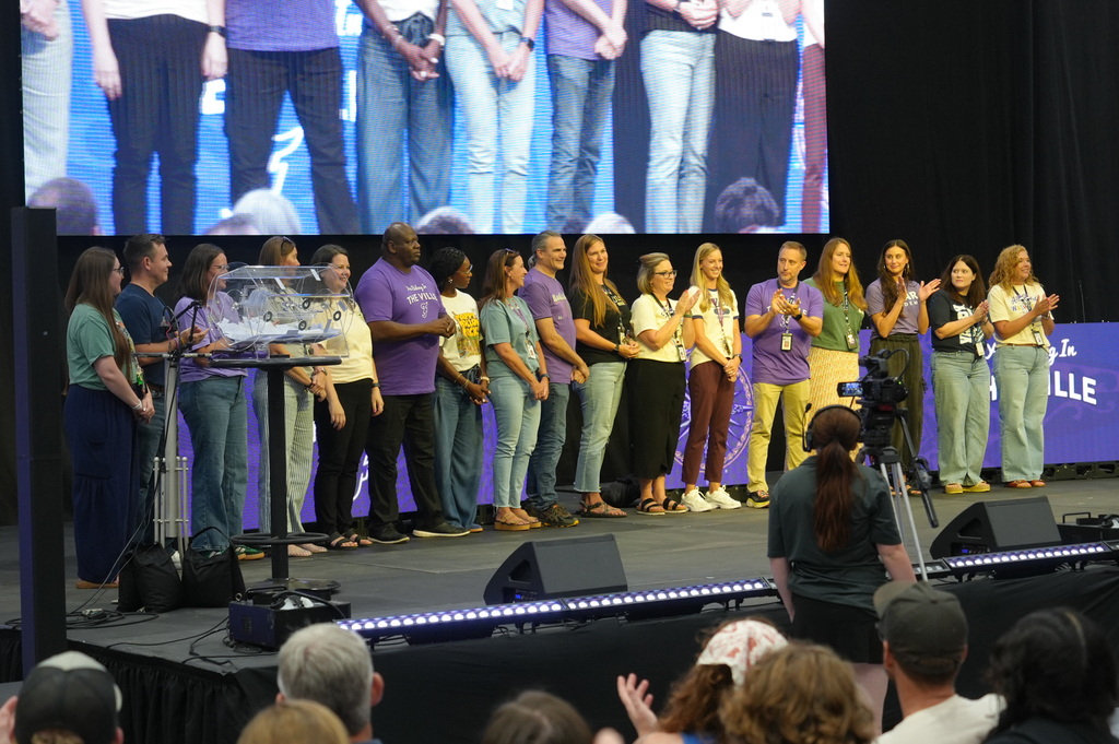 A group of people on stage, some clapping, a large screen behind them, and an audience in front.