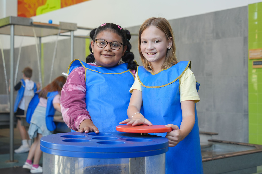 Two girls in blue aprons stand with a blue recycling bin and red disk. A playground and other children are in the background.