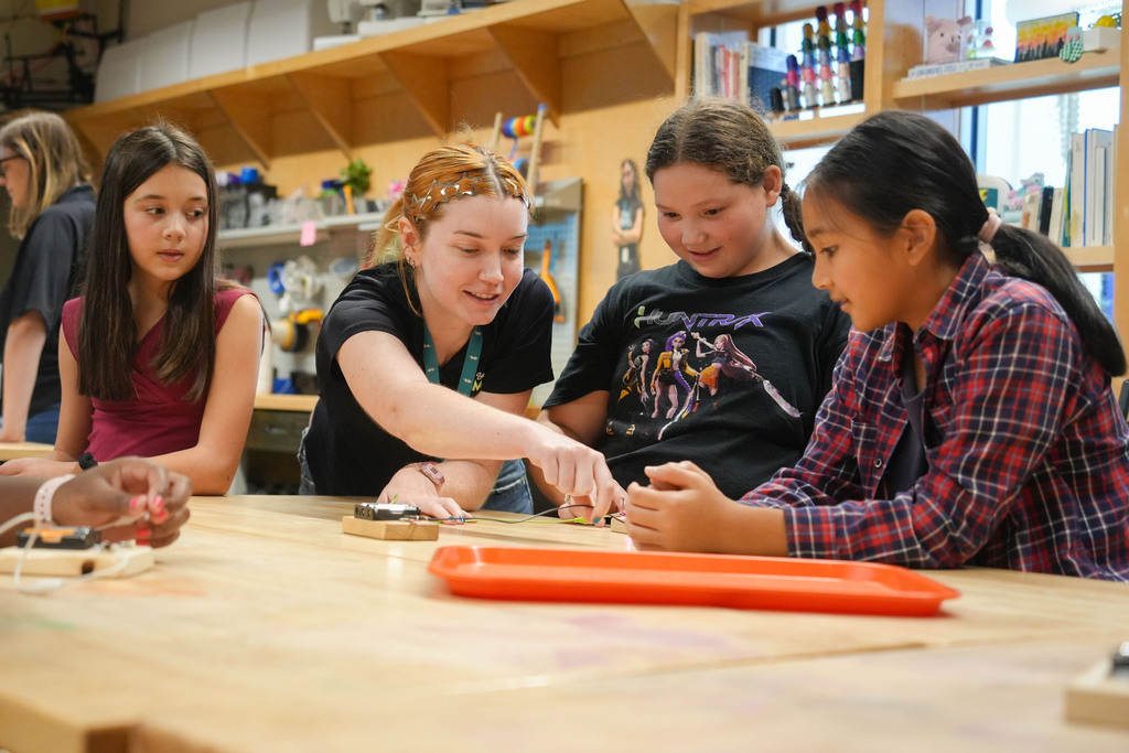 Five girls in a classroom are working together on a project on a table. A woman is guiding two of the girls.