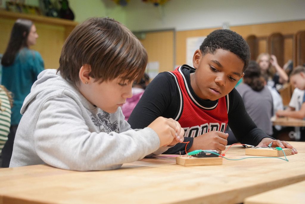 Two children seated at a wooden table, working on a small electronics project. They are using wires and small tools.