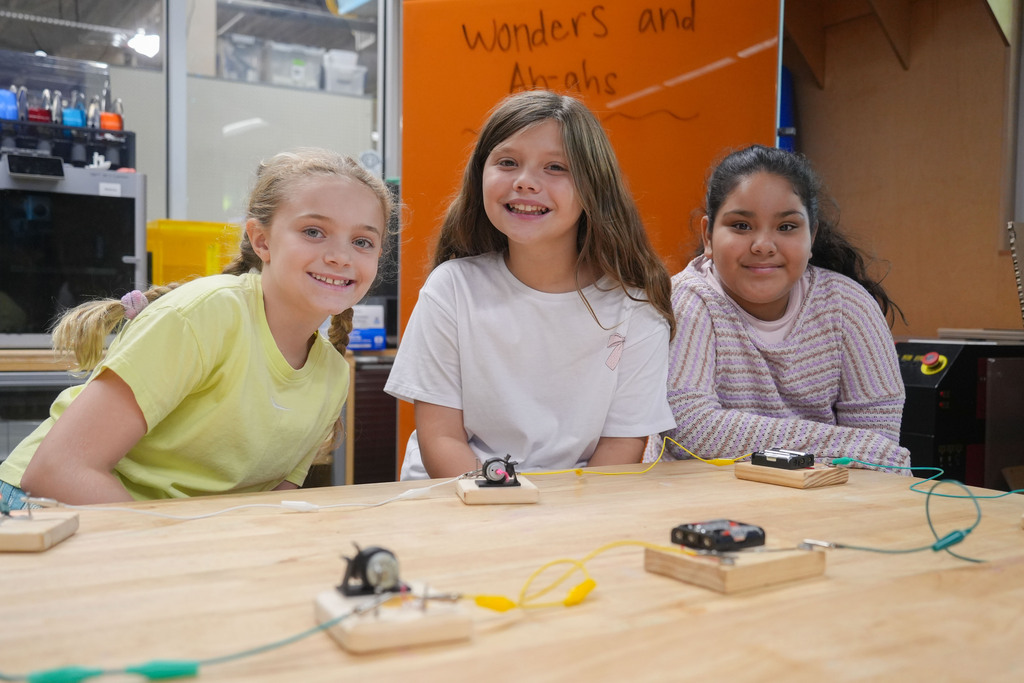 Three young girls work together at a table, focused on a science project with wires and small devices.