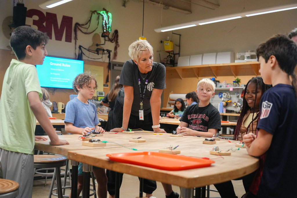 A woman guides a group of children in a classroom, standing at a table with electronic components.