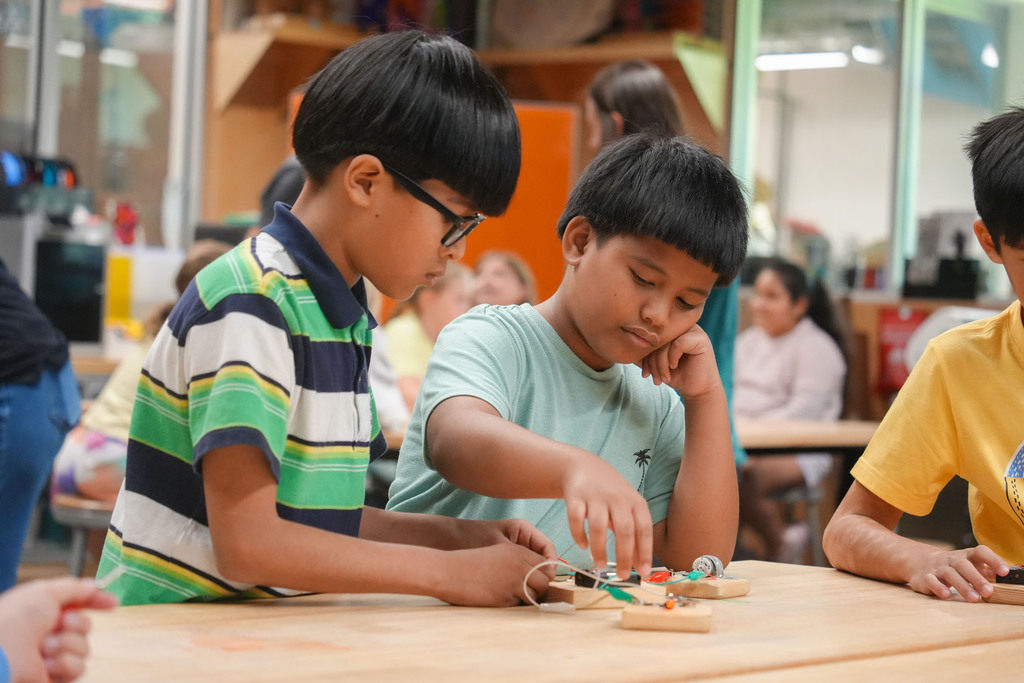 Three children focused on a table with a small circuit board, one wearing glasses, another with his eyes closed.