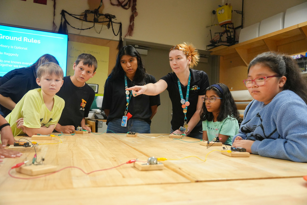 Children and adults in a classroom. An instructor points to a circuit on a table.