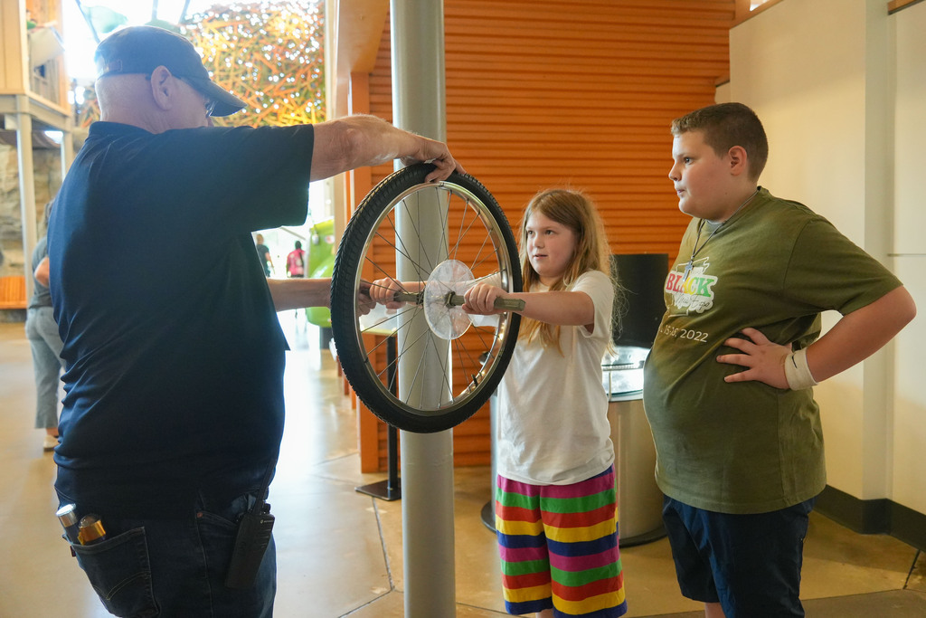 A man and a girl examine a bicycle wheel attached to a pole, while a boy looks on.