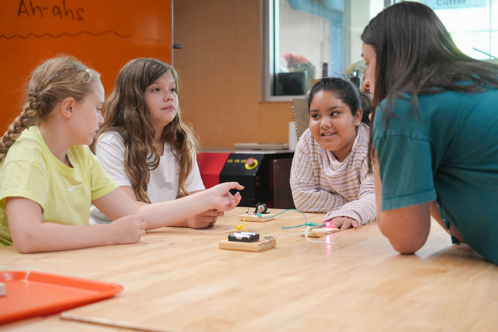 Four children seated around a table. One child has braided hair, and a woman stands next to her.