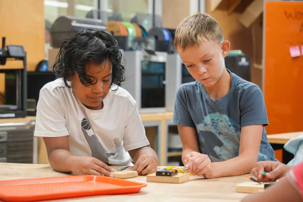 Two children focus on a circuit board at a table, surrounded by electronic equipment.