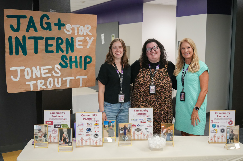 Three women stand behind a table displaying pamphlets and books; a sign reads "JAG+STAR INTERNET SHIP JONES TROUT".