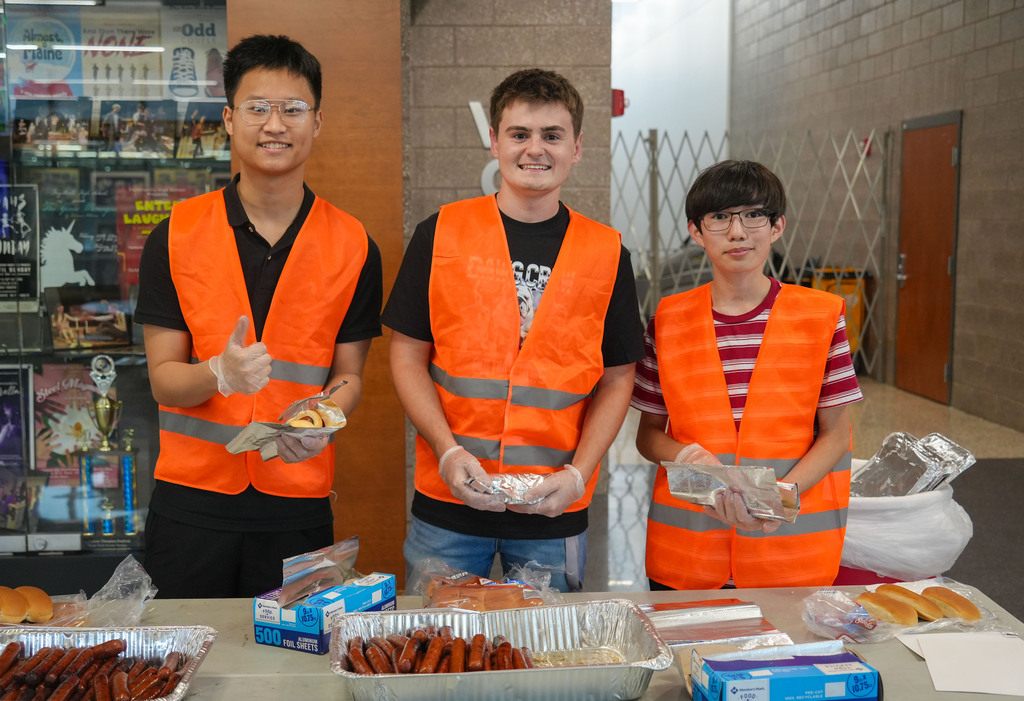 Three people in orange vests stand by a table with food items, against a backdrop of a store.