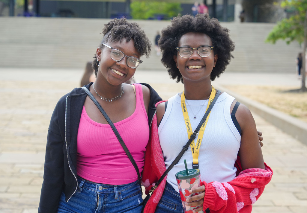 Two women standing together, both smiling and holding drinks, wearing glasses and different colored tops. Behind them is a building with a wall and trees.