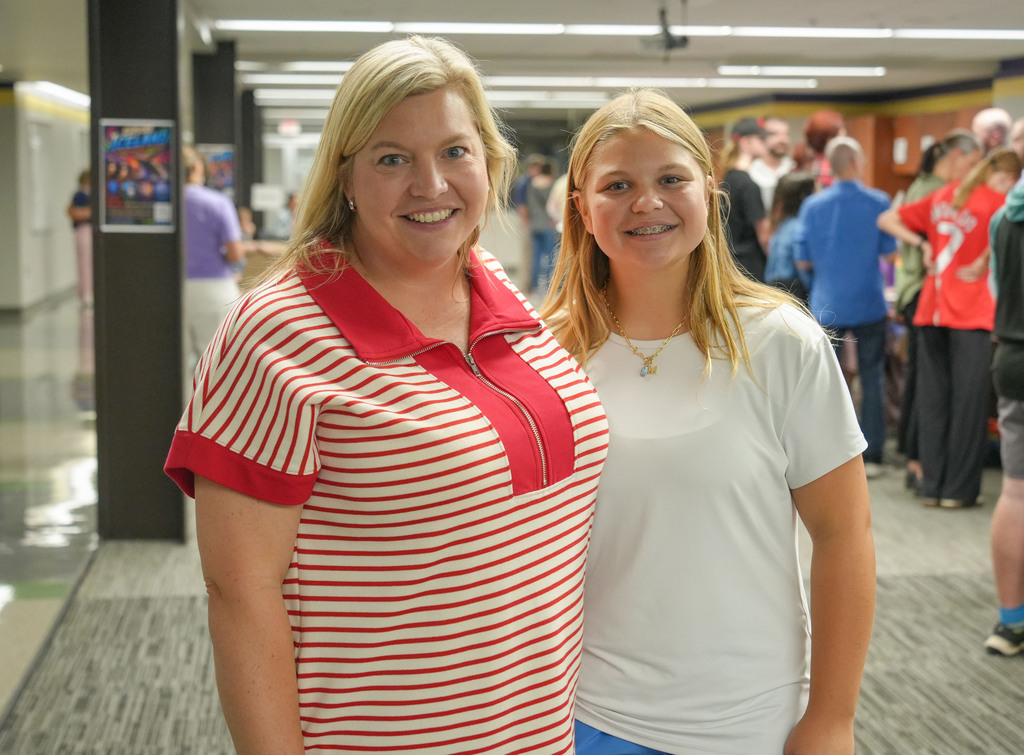 Two women stand next to each other in a hallway with several people in the background.