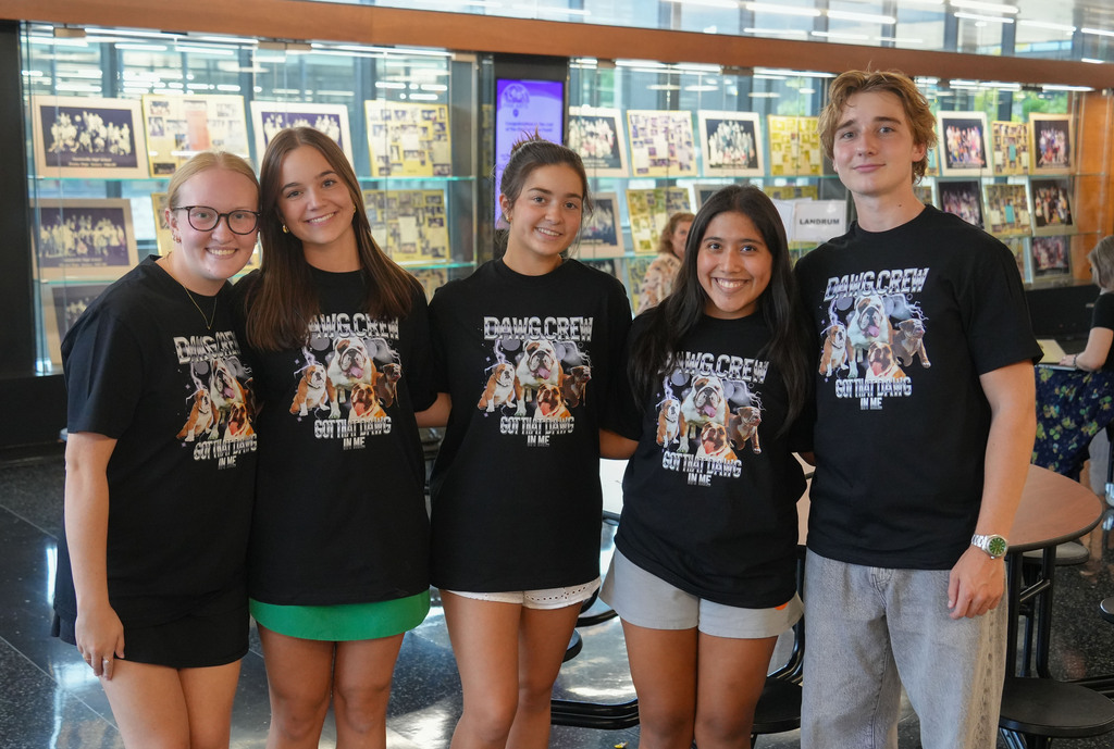 Five young people wearing matching black T-shirts stand together. One wears glasses, and they are in front of a display case.