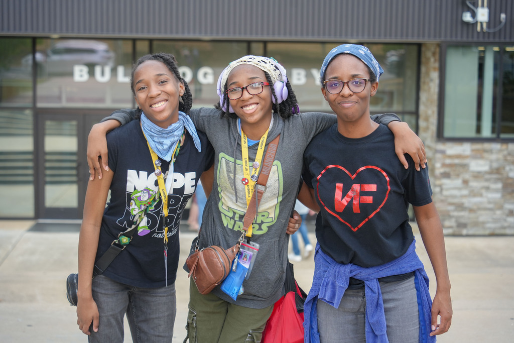 Three young women stand together in front of a building. One wears a scarf, another wears a backpack.