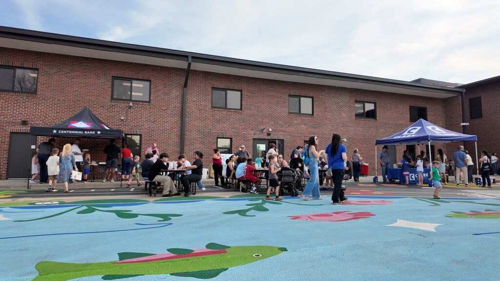 People gather in front of a building with a painted mural, under tents. Some are seated at tables.