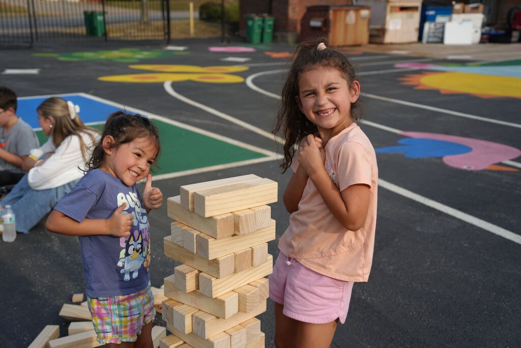 Two girls playing with a wooden block tower on a playground. A colorful painted background and other children in the distance.