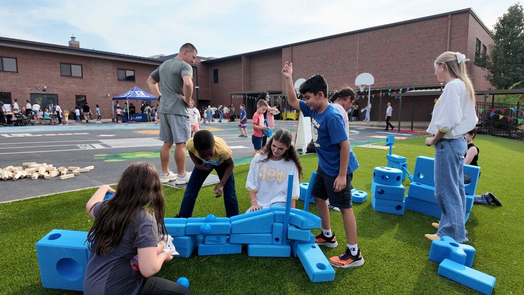 Children and adults play with blue blocks on a green turf. Behind them, a building with many windows stands.