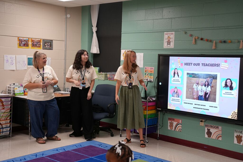 Three women in front of a large screen in a classroom with a rug on the floor.