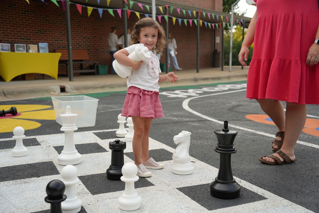 Child wearing a white shirt and pink shorts stands at a giant chessboard on a sidewalk.