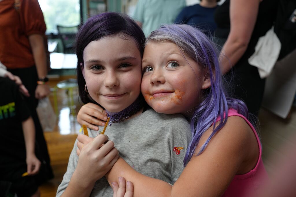 Two girls with purple hair smile and hug. One wears gray, the other pink. Blurry figures, including adults, in background.