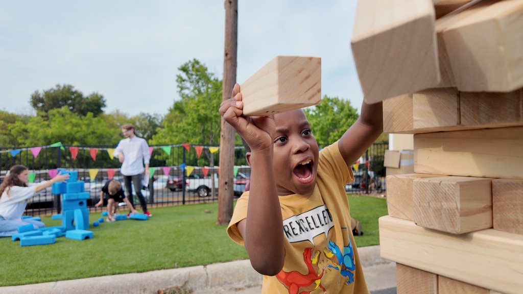 Child holds up wooden block, shouts, and laughs. Behind, a playground with other children and adults.